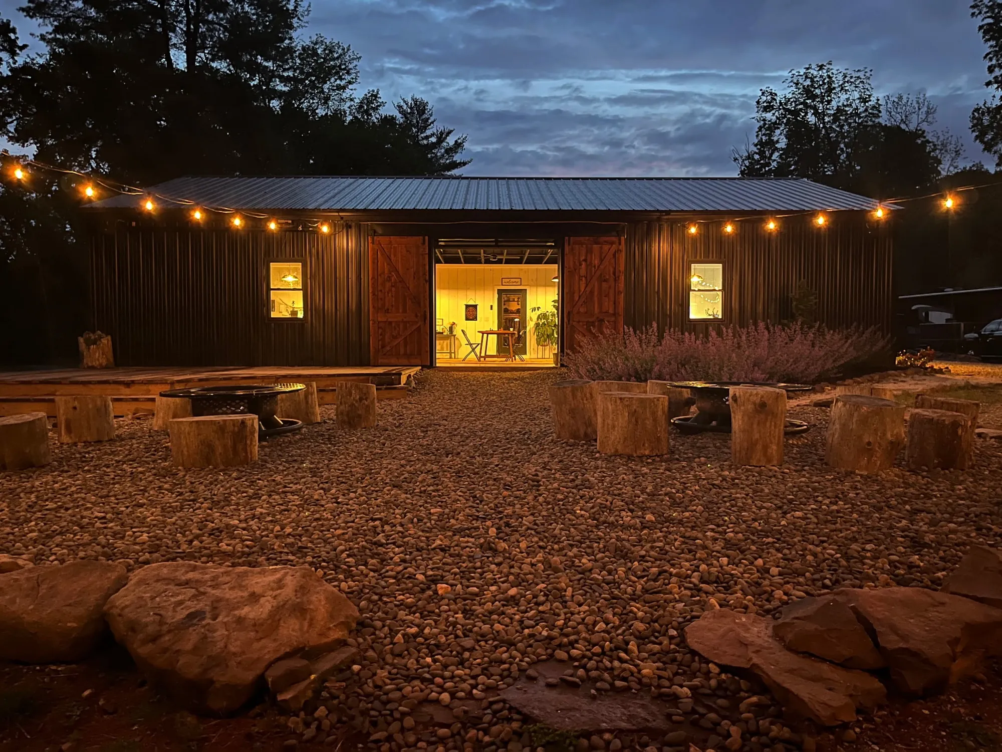 The fire circle at night, ringed with log stools under string lights in front of the rec room