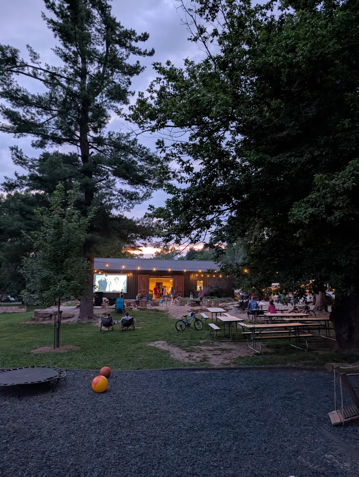 Campers in chairs watching a movie on the outdoor screen at dusk, the rec room lit up behind them
