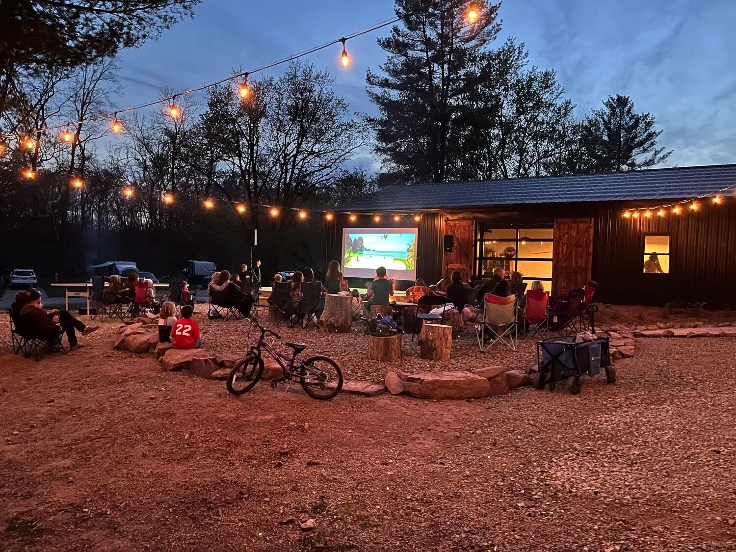 Campers around the fire circle for an outdoor movie under string lights