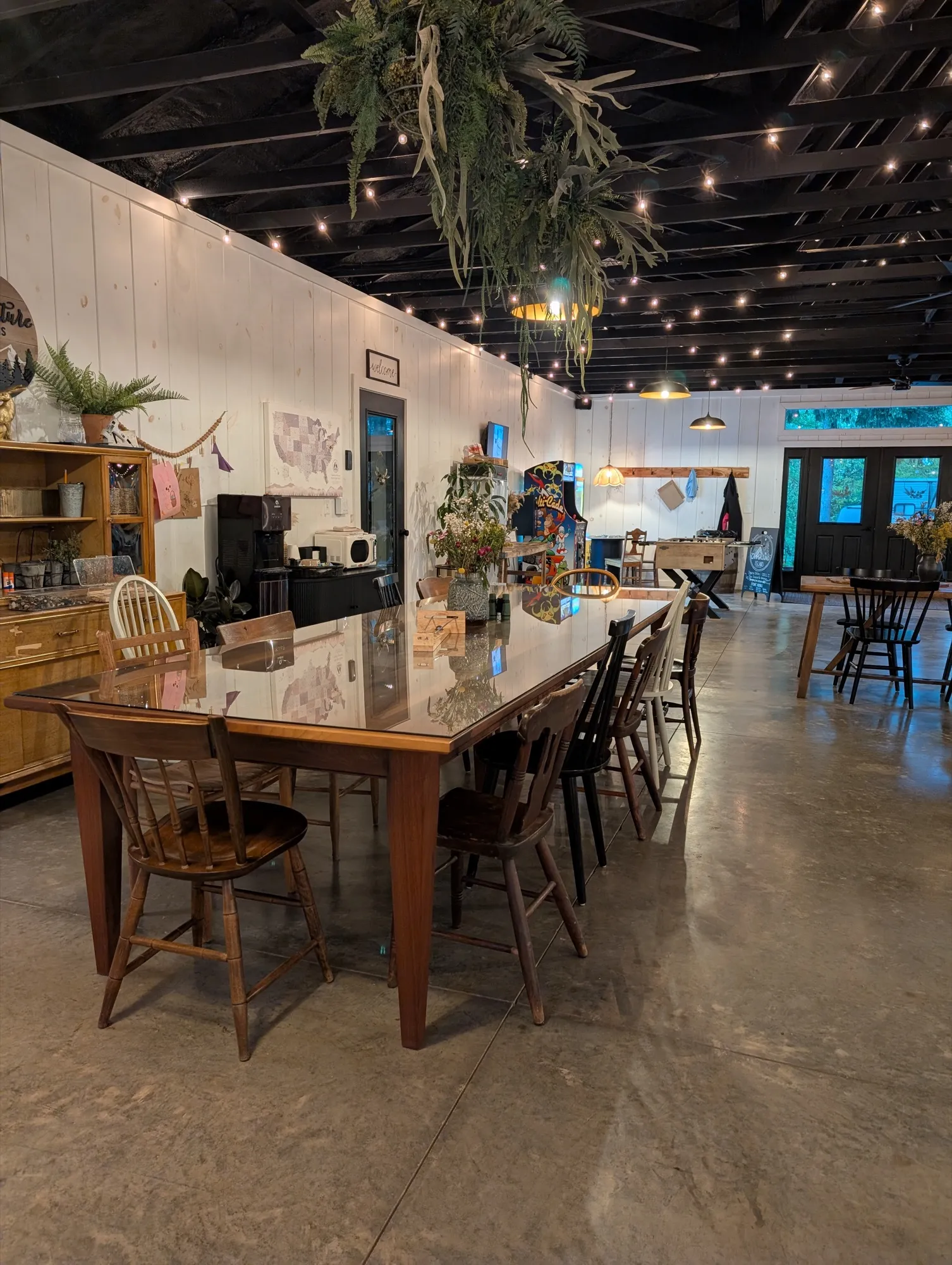 The rec room's long farm table set with mismatched wooden chairs under string lights