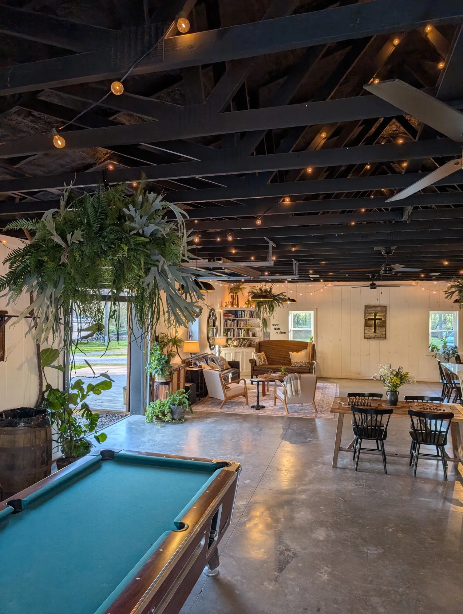 The rec room under string lights, with a pool table in front and dining tables beyond