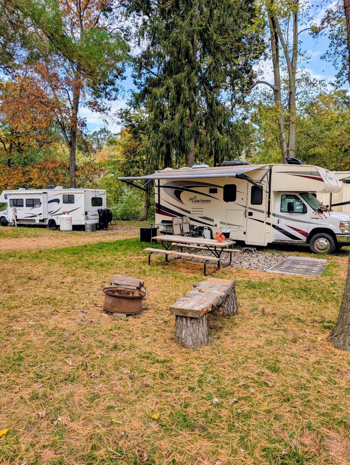 A motorhome and a travel trailer on their sites, with a picnic table, fire ring and log bench